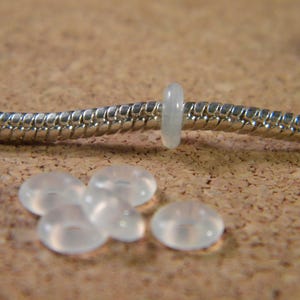 Peut inclure: Gros plan sur un bracelet en chaîne serpent argenté avec une perle en plastique transparente. Cinq autres perles en plastique transparentes se trouvent sur la surface sous le bracelet.