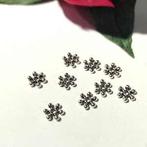 May include: Silver metal flower-shaped beads with six points. The beads are scattered on a white surface.