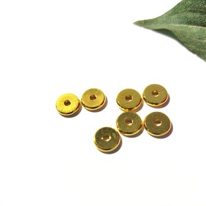 May include: A group of seven gold-colored metal beads, each with a round shape and a hole in the center. The beads are scattered on a white surface.