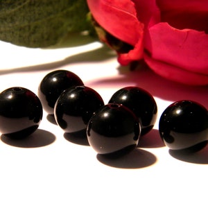 May include: Six black round beads, possibly made of onyx or obsidian, are arranged in a row on a white surface. The beads are smooth and shiny, reflecting light.