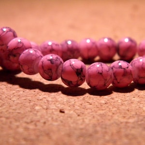 Peut inclure: Un collier de perles rose et noir avec un motif marbré. Les perles sont rondes et ont une finition brillante.