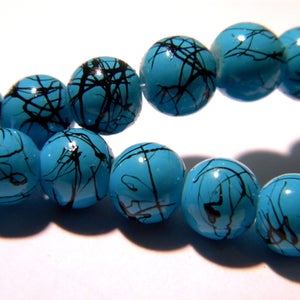 May include: A strand of turquoise beads with black, abstract paint strokes. The round beads are a bright, glossy blue-green color. These beads could be used for jewelry making or other craft projects.