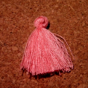 Puede incluir: Un borlón rosa con un nudo en la parte superior. El borlón está hecho de hilo suave y esponjoso y mide aproximadamente 2,5 cm de largo.