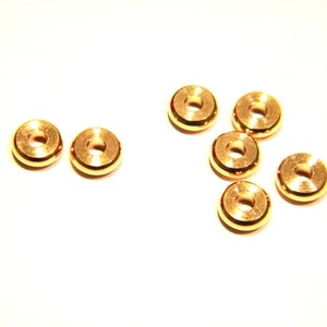 May include: Several small, gold-colored metal rings with a circular design and a hole in the center are scattered on a white surface. The rings appear to be made of a shiny, metallic material.