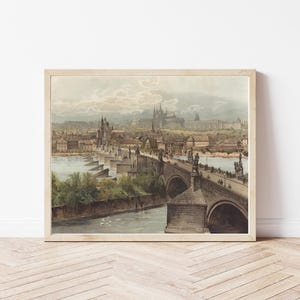 Puede incluir: Una impresión vintage enmarcada de una ciudad europea, con un puente de piedra sobre un río. La obra de arte muestra un paisaje urbano detallado con edificios y un cielo nublado, en tonos apagados.