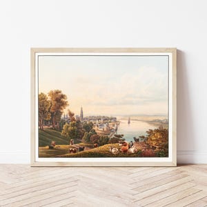 Op de afbeelding: Ingelijste landschapsprint met een gedetailleerd uitzicht op een Europese stad langs een rivier. Het kunstwerk toont een levendig kleurenpalet met aandacht voor de architectuur, bomen en figuren op de voorgrond. De print zit in een lichtgekleurde houten lijst.