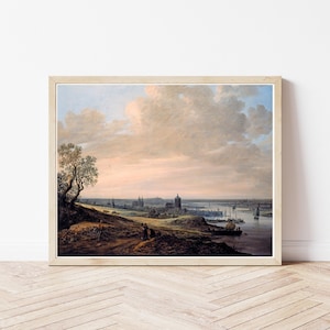 Op de afbeelding: Ingelijste kunstafdruk met een gedetailleerde weergave van een stad aan een rivier onder een bewolkte lucht. Het kunstwerk toont een historische scène met gebouwen, boten en figuren op een heuvel. Het kleurenpalet omvat gedempte tinten bruin, blauw en roze.