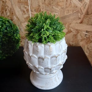 Könnte beinhalten: Ein weißer, dekorativer Pflanztopf mit abgenutztem Goldbesatz, gefüllt mit leuchtend grünem Kunstblattwerk. Der Pflanztopf hat ein einzigartiges, strukturiertes Design und steht auf einem kleinen Sockel. Eine grüne Topiarkugel ist im Hintergrund zu sehen.