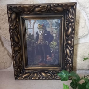 Puede incluir: Un cuadro enmarcado que muestra una rana elegante con traje, en un jardín exuberante. El marco dorado y marrón tiene detalles ornamentales. La obra de arte tiene un estilo vintage, con la rana usando chaleco y sombrero de copa.