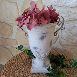 Peut inclure: Un vase blanc vieilli avec des accents gris foncé, rempli de fleurs roses. Le vase a des poignées décoratives et repose sur une table ronde tressée. Le lierre vert ajoute une touche de couleur naturelle.