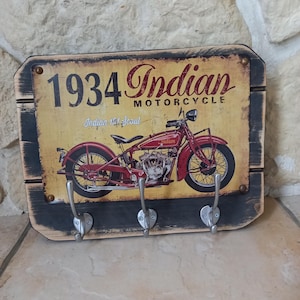 Può includere: Portachiavi da parete in legno con un design di moto vintage. L'insegna recita "1934 Indian Motorcycle" con una grafica di moto rossa. Include tre ganci in metallo argentato per appendere chiavi o altri piccoli oggetti. Il legno ha un aspetto rustico.