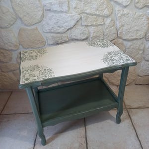 Peut inclure: Petite table d'appoint de style ancien, avec un plateau blanc et une étagère inférieure. La table a un cadre et des pieds vert sauge avec un motif floral décoratif dans les coins du plateau. La table mesure environ 61 cm de long, 41 cm de large et 61 cm de haut.