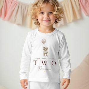 Op de afbeelding: Een wit setje met een shirt met lange mouwen en een broek, voorzien van een afbeelding van een teddybeer die een ballon vasthoudt. De tekst "WHEN I WAKE I'LL BE TWO" staat in bruin op de voorkant, gevolgd door de naam "Roman" in cursief.