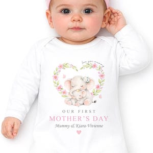 Puede incluir: Un bebé con un body blanco y una diadema rosa con lazo. El body presenta un diseño floral en forma de corazón con dos elefantes y el texto "OUR FIRST MOTHER'S DAY". El texto "Mummy & Kiara-Vivienne" también está impreso en el body.