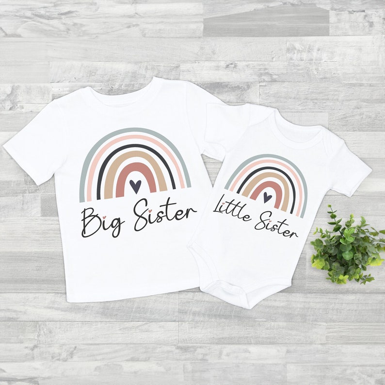 Puede incluir: Conjunto de camiseta y body blancos con un dise&ntilde;o de arco&iacute;ris en colores apagados. La camiseta dice "Big Sister" y el body "little Sister". Un coraz&oacute;n est&aacute; en el centro del arco&iacute;ris.