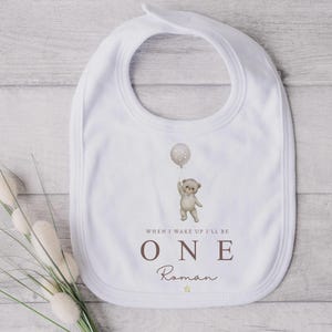 Op de afbeelding: Een wit slabbetje met een centrale illustratie van een lichtbruine teddybeer die een ballon met stippen vasthoudt. Onder de beer staat de tekst: WHEN I WAKE UP I'LL BE ONE, gevolgd door de naam Roman en een klein geel sterretje.