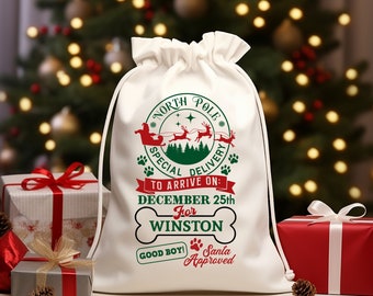 Op de afbeelding: Een witte kerstcadeauzak met trekkoord en feestelijke afbeeldingen. De tas heeft een Noordpool-ontwerp met de kerstman en rendieren, de tekst "Special Delivery" en "To Arrive On: December 25th For Winston." Inclusief een hondenbot en "Good Boy! Santa Approved."