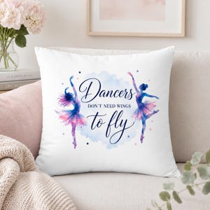 A white square pillow with two watercolor ballerinas and the text "Dancers don't need wings to fly" is on a light-colored sofa with a pink blanket and a vase of flowers.