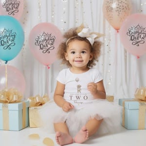 Op de afbeelding: Een wit rompertje met korte mouwen met een afbeelding van een teddybeer die een ballon vasthoudt en de tekst "WHEN I WAKE UP I'LL BE TWO Daisy". Het rompertje is gecombineerd met een witte tule tutu-rok en een witte haarstrik met gouden glitteraccenten.