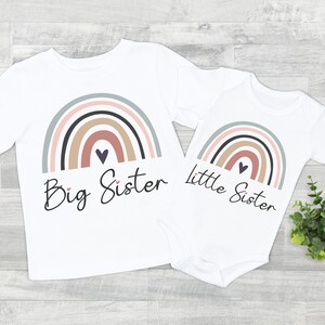 Puede incluir: Conjunto de camiseta y body blancos con dise&ntilde;o de arco&iacute;ris en colores apagados. La camiseta dice "Big Sister" y el body "Little Sister" en fuente cursiva. Un coraz&oacute;n est&aacute; debajo del arco&iacute;ris.