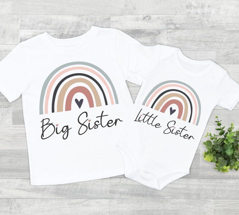 Puede incluir: Conjunto de camiseta y body blancos con dise&ntilde;o de arco&iacute;ris en colores apagados. La camiseta dice "Big Sister" y el body "Little Sister" en fuente cursiva. Un coraz&oacute;n est&aacute; debajo del arco&iacute;ris.
