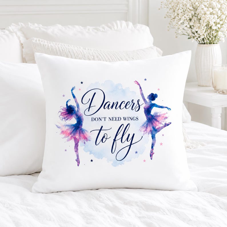 A white square pillow with two watercolor ballerinas and the text "Dancers don't need wings to fly" is centered on a white bed with a vase of white flowers.