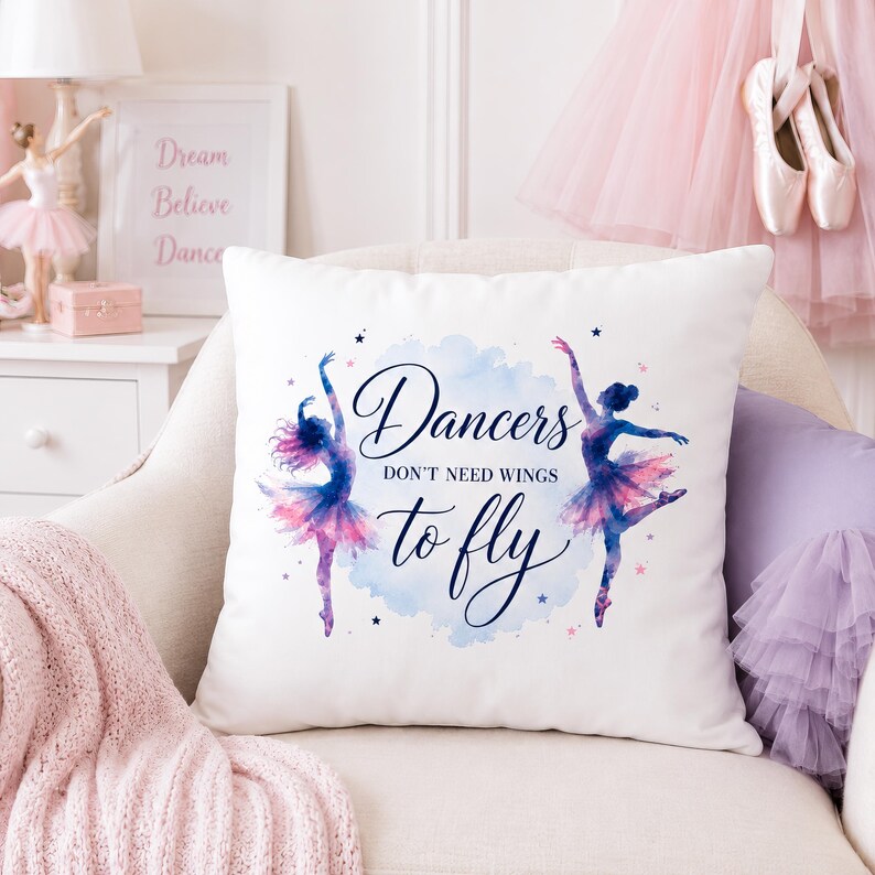 A white square pillow with two watercolor ballerinas and the text "Dancers don't need wings to fly" rests on a white chair with pink ballet decor in the background.