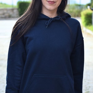 Puede incluir: Una sudadera con capucha azul marino con capucha con cord&oacute;n y bolsillo delantero. La sudadera est&aacute; hecha de un material suave y tiene un ajuste relajado. La imagen muestra la parte superior del cuerpo de una persona que lleva la sudadera con capucha.