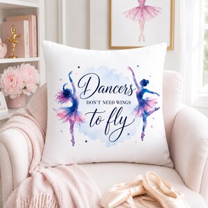 A white square pillow with two watercolor ballerinas and the text "Dancers don't need wings to fly" is on a pink armchair with ballet slippers and pink floral decor.