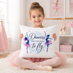A young girl in a pink tutu holds a white square pillow with two watercolor ballerinas and the text "Dancers don't need wings to fly."