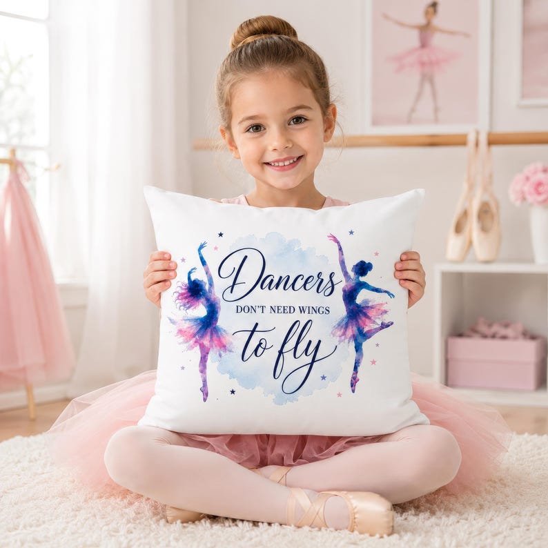 A young girl in a pink tutu holds a white square pillow with two watercolor ballerinas and the text "Dancers don't need wings to fly."