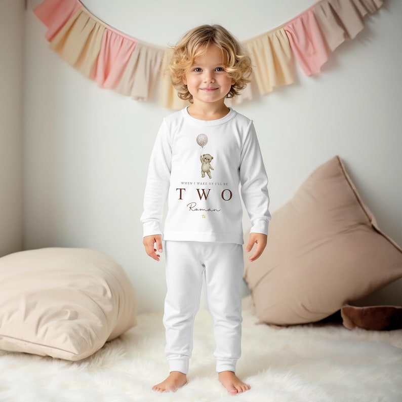 Op de afbeelding: Een wit setje met een shirt met lange mouwen en een broek, voorzien van een afbeelding van een teddybeer die een ballon vasthoudt. De tekst "WHEN I WAKE I'LL BE TWO" staat in bruin op de voorkant, gevolgd door de naam "Roman" in cursief.
