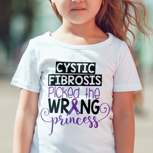 Puede incluir: Una camiseta blanca con una cinta morada y el texto "CYSTIC FIBROSIS picked the WRONG princess".