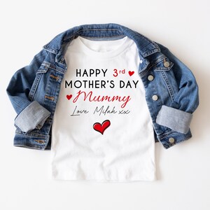 May include: White t-shirt with the text "HAPPY 3rd MOTHER'S DAY Mummy Love Milah xx" in red and black. A red heart graphic is below the text. The t-shirt is paired with a blue denim jacket.
