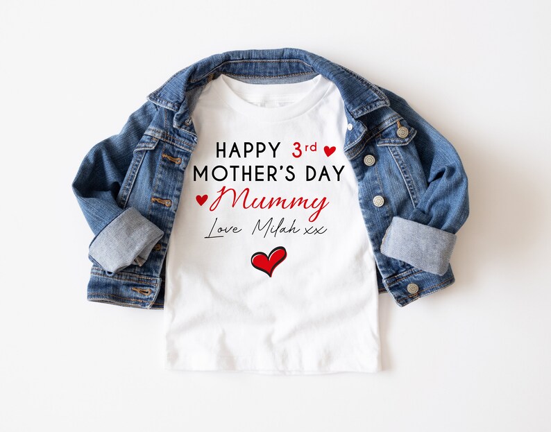 May include: White t-shirt with the text "HAPPY 3rd MOTHER'S DAY Mummy Love Milah xx" in red and black. A red heart graphic is below the text. The t-shirt is paired with a blue denim jacket.