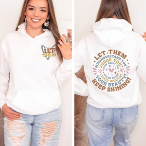Puede incluir: Sudadera con capucha blanca con bolsillo delantero y capucha. La parte delantera presenta el texto "LET THEM" en un dise&ntilde;o en forma de coraz&oacute;n. La parte trasera muestra un dise&ntilde;o circular con el texto "LET THEM MISUNDERSTAND YOU...KEEP SHINING!"