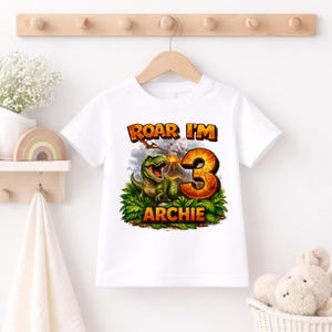 Personalised dinosaur birthday shirt hanging on wooden hanger, custom name Archie age 3, white kids T-shirt, T-Rex volcano design