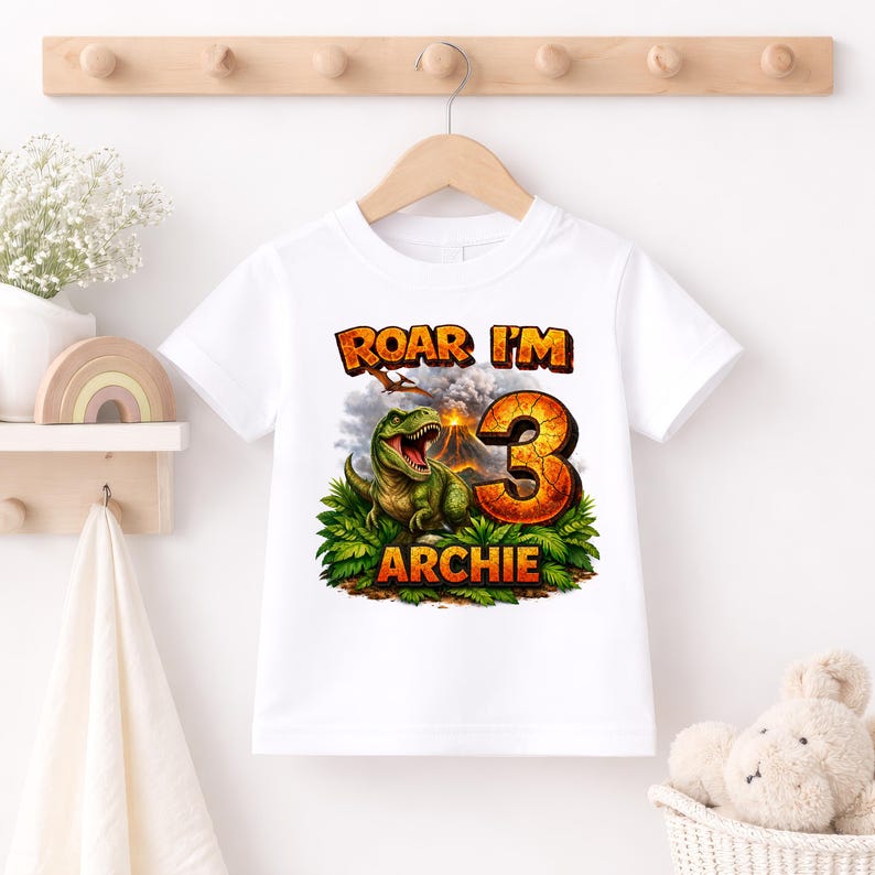 Personalised dinosaur birthday shirt hanging on wooden hanger, custom name Archie age 3, white kids T-shirt, T-Rex volcano design