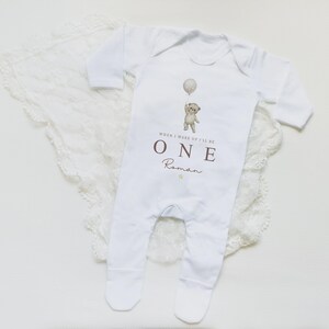 Op de afbeelding: Een witte baby-onesie met lange mouwen en voetjes, voorzien van een afbeelding van een teddybeer die een ballon vasthoudt. De tekst luidt "WHEN I WAKE UP I'LL BE ONE" in bruine hoofdletters, gevolgd door de naam "Roman" in bruin handschrift.