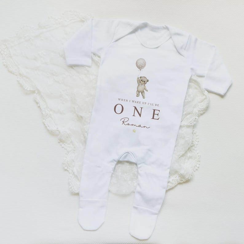Op de afbeelding: Een witte baby-onesie met lange mouwen en voetjes, voorzien van een afbeelding van een teddybeer die een ballon vasthoudt. De tekst luidt "WHEN I WAKE UP I'LL BE ONE" in bruine hoofdletters, gevolgd door de naam "Roman" in bruin handschrift.