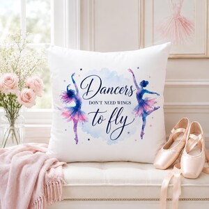 A white square pillow with two watercolor ballerinas and the text "Dancers don't need wings to fly" is on a white bench with ballet slippers and a pink throw.