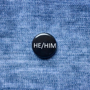 May include: A black button badge with the white text "HE/HIM". The badge is positioned on a blue denim fabric, providing a contrast between the dark badge and the lighter material.