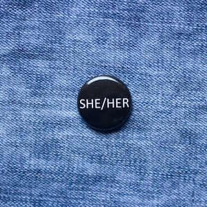 May include: A black button badge with the white text "SHE/HER". The badge is positioned on a blue denim fabric background.