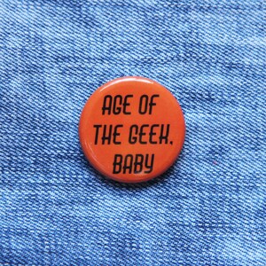 May include: Orange button with the text "Age of the Geek. Baby" on a blue denim background.