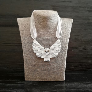 Op de afbeelding: Een witte kralenketting met een uil, zwarte ogen en een wit lint. De ketting wordt getoond op een grijze mannequin.