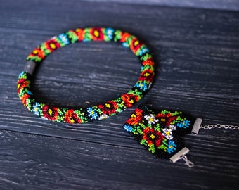May include: A beaded necklace and bracelet set. The necklace is a thick, round design with a magnetic clasp. The bracelet has a silver chain and clasp. Both pieces feature a floral pattern in red, yellow, green, and blue beads.