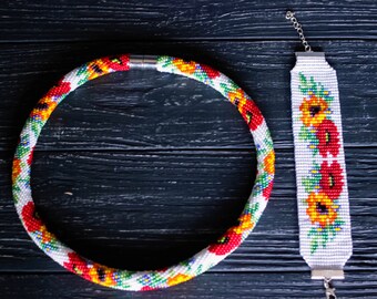 May include: A beaded necklace and bracelet set. The necklace is a white base with a pattern of red, yellow, and green beads. The bracelet is white with a floral pattern of red and yellow flowers. The set is displayed on a dark wood surface.