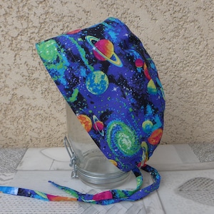 May include: A blue surgical cap with a colourful space-themed print featuring planets, stars, and galaxies. The cap has long ties that can be tied at the back of the neck.