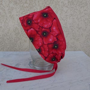 Può includere: Una cuffia in tessuto caratterizzata da un vivace motivo a fiori di papavero rosso con centri scuri. Il copricapo ha una forma arrotondata e include due lunghi lacci in tessuto rosso tinta unita che partono dal bordo inferiore per fissarlo.