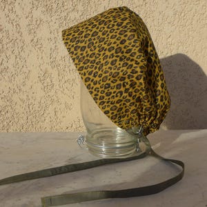 Pode incluir: Uma touca cirúrgica com estampa de leopardo em tons de ouro e marrom, repousando em um pote de vidro. A touca tem um design franzido e é amarrada com laços de tecido verde-oliva. O fundo é uma parede bege texturizada.